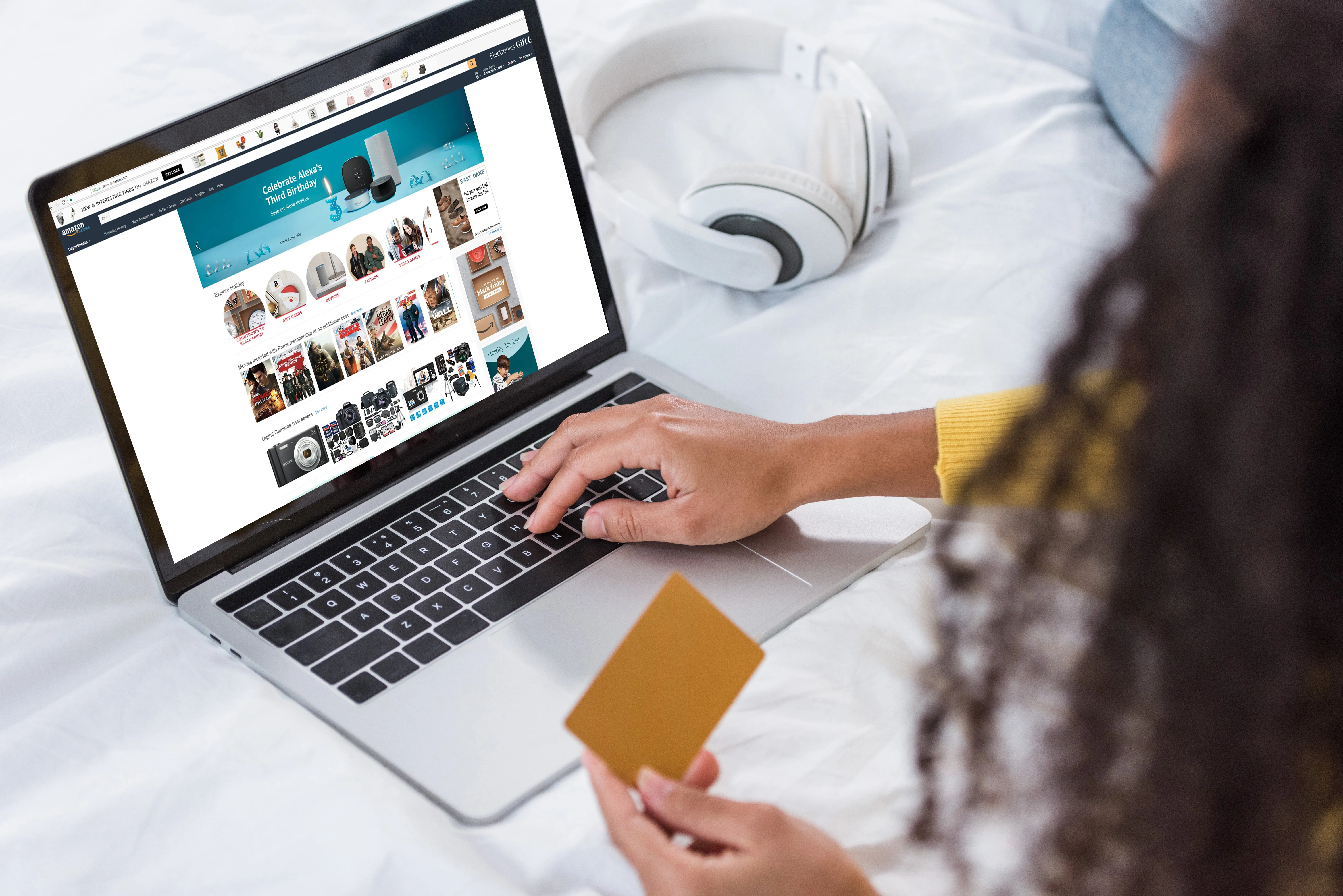 Eine Frau mit langen dunklen Haaren und einem gelben Oberteil sieht sich Elektrogeräte auf einem Laptop in einem Onlineshop an. Sie hält eine Kreditkarte in der Hand.